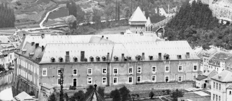 Vue ancienne en noir et blanc d’un grand bâtiment en pierre à toit incliné, avec de nombreuses fenêtres en façade et des lucarnes sur le toit. À l’arrière-plan, un paysage vallonné, une forêt dense et un pont menant à une structure de style médiéval avec une tourelle. (L'ANCIENNE CASERNE DE CAVALERIE (PFAFFENTHAL)