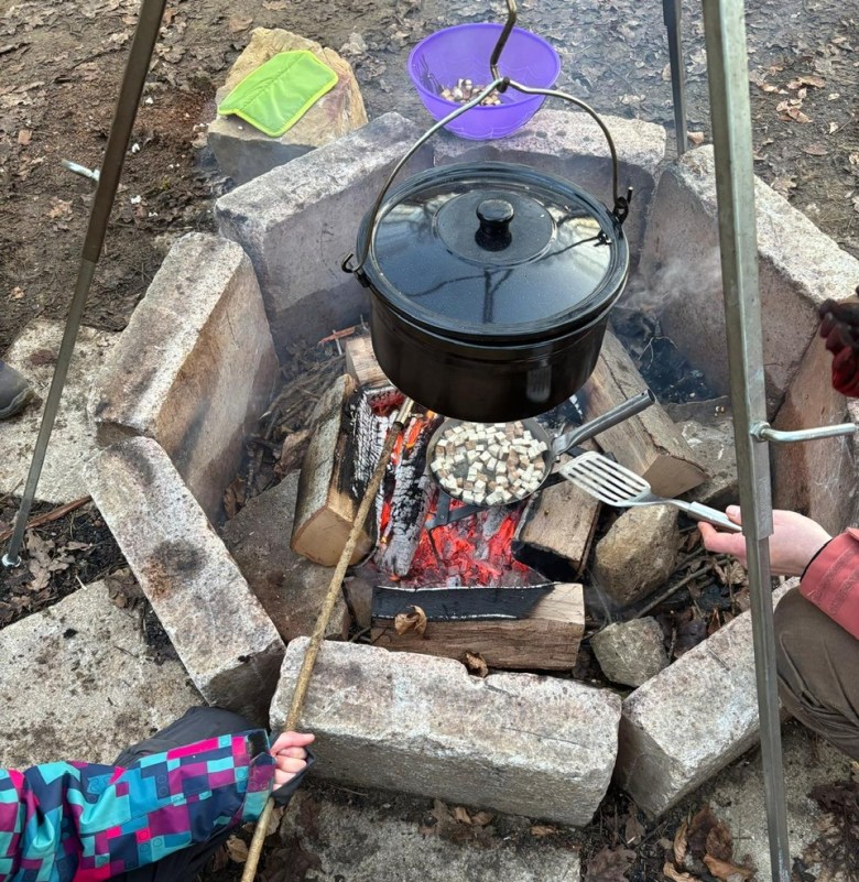 Des personnes cuisinent en plein air autour d’un feu de camp entouré de blocs de pierre. Une grande marmite noire est suspendue au-dessus du feu, tandis qu’une poêle contenant des petits morceaux de nourriture est maintenue au-dessus des braises. Un enfant en manteau coloré tient un bâton, et un adulte utilise une spatule pour cuisiner.
